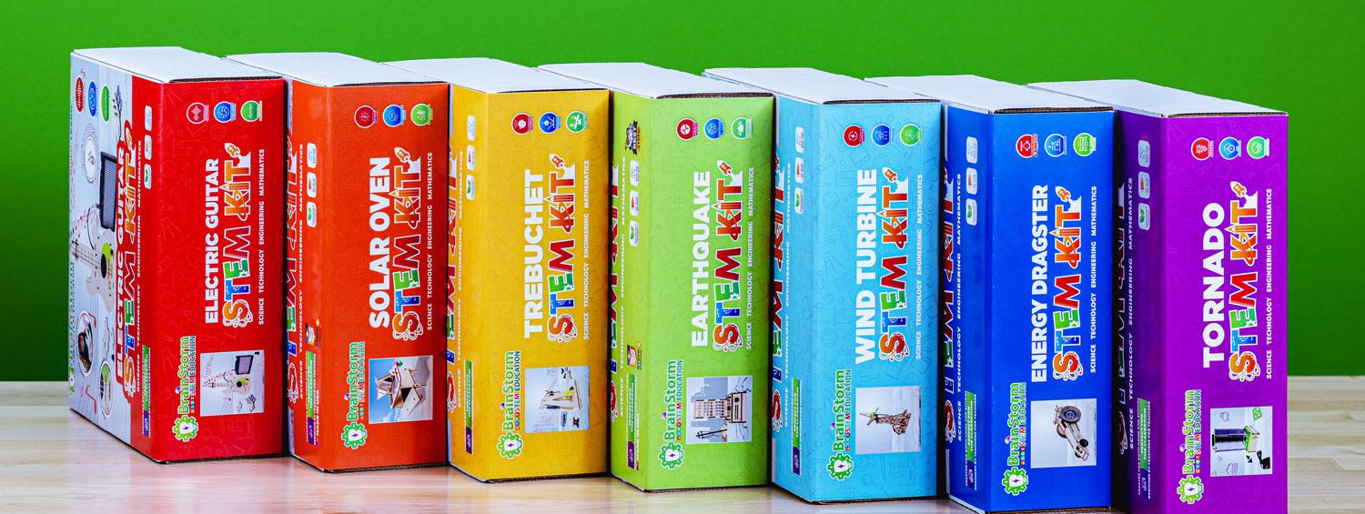 A row of seven BrainStorm individual STEM kit boxes, including Electric Guitar, Solar Oven, Trebuchet, Earthquake, Wind Turbine, Energy Dragster, and Tornado, standing on a table against a green background.